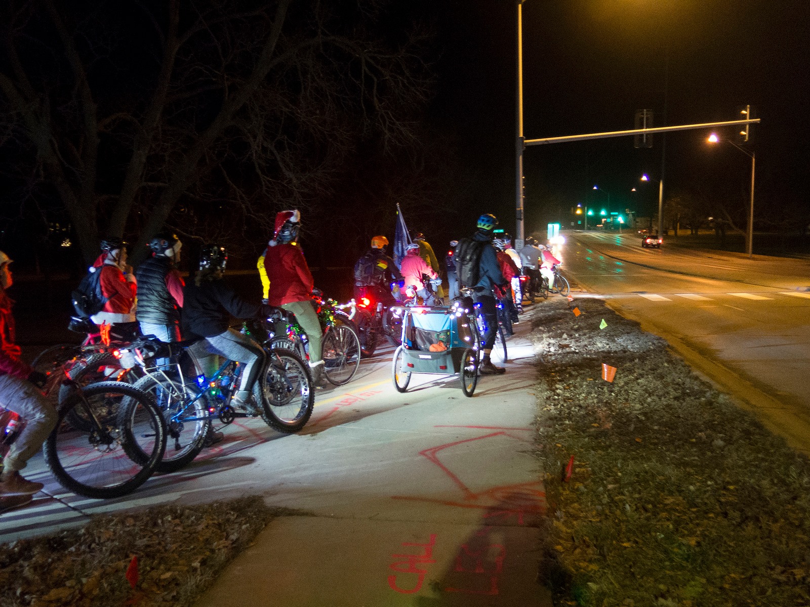 Group of bicycle riders, some dressed as santas, wait to cross a street in the dark.